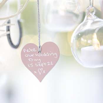 A heart-shaped piece of wood hangs between various-sized glass candle holders. The holders are also suspended. Upon the wooden heart is a handwritten message reading 'Our Wedding Day 15 Sept 22'.