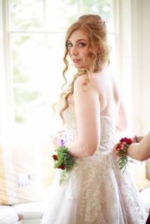 A%20bride%20stood%20in%20front%20of%20a%20large%20window.%20Her%20dress%20is%20being%20fastened%20as%20she%20glances%20at%20the%20camera.